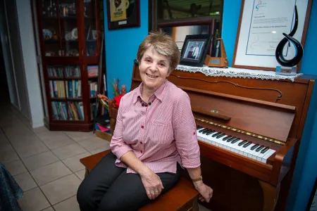 A smiling woman sits at a piano in her home, wearing a pink button-up shirt and dark pants. Behind her on the piano are framed photos, certificates, and music-related memorabilia, with a bookshelf visible in the background.
