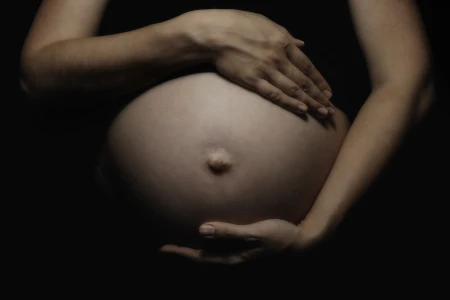 Close-up of a pregnant belly with two hands gently cradling it, one resting above and one below, against a dark background.