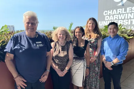 Five people stand together smiling on a sunny balcony at Charles Darwin University. The group includes three women and two men, dressed in smart casual attire, with lush green plants and a clear blue sky in the background. The CDU logo is visible on the building wall behind them.