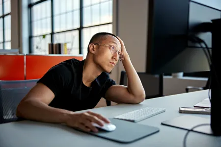 A person wearing glasses and a black t-shirt sits at a desk in front of a computer, resting their head on one hand while using a mouse with the other. They appear tired or frustrated, gazing off into the distance in a modern office with large windows and natural light.