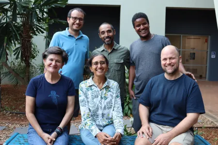 six people, 3 seated,  standing behind them, posing for this photo, with a building and palm tree behind them