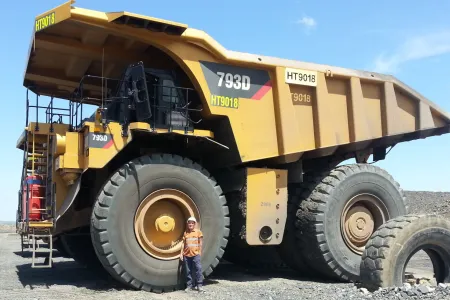 Very large mine truck with person standing by front wheel, looking tiny in comparison to truck size