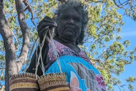 Associate Professor Elaine Ḻäwurrpa Maypilama holding woven dilly bags on country