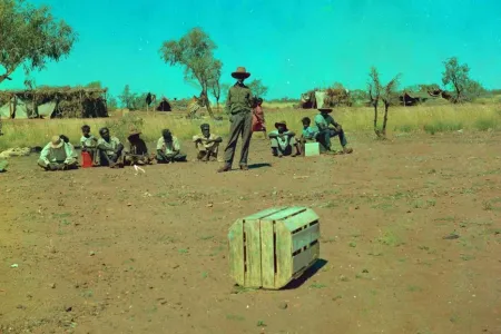 Photo containg wooden barrel in the fron with a group of Indigenous people sitting far back while Don McLeod (whiteman) is standing