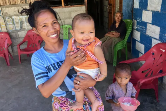 A woman holding a small child with another child standing beside her and an old woman sitting in the background
