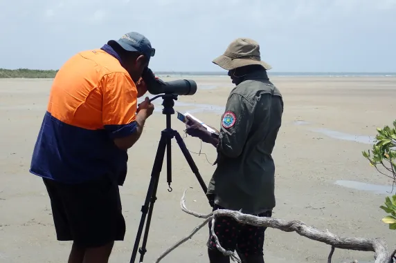 two men on a beach, one looking through a telescope, one writing in a notebook