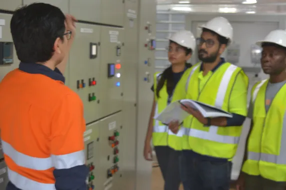 Three people in yellow hi-vis vests and facing camera and one person in orange hi-vis with back to camera looking at grey instrument panel