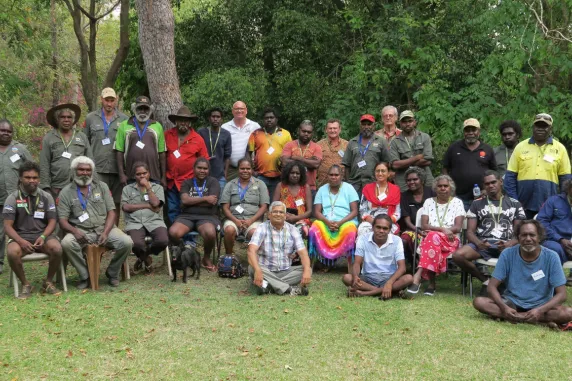 Group photo of about 35 people, some sitting on chairs, some sitting on the ground, and some standing. Tree trunk and green leafy bushes in background