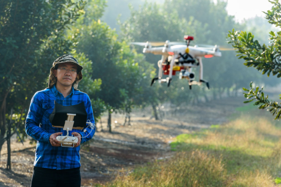 person wearing hat and blue checked shirt standing in field with trees holding drone control unit, drone in flight in foreground