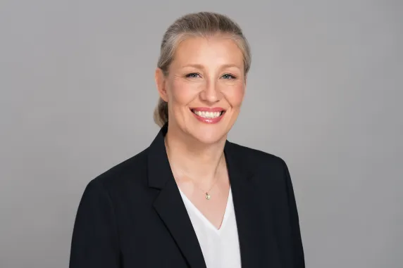 Woman with black jacket, blonde hair, blue eyes, smiling towards camera