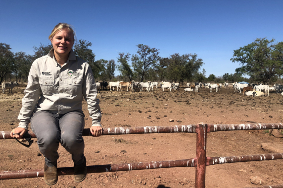 Beth Penrose sitting on a rusty steel fence with bare earth, cattle, and trees in the background