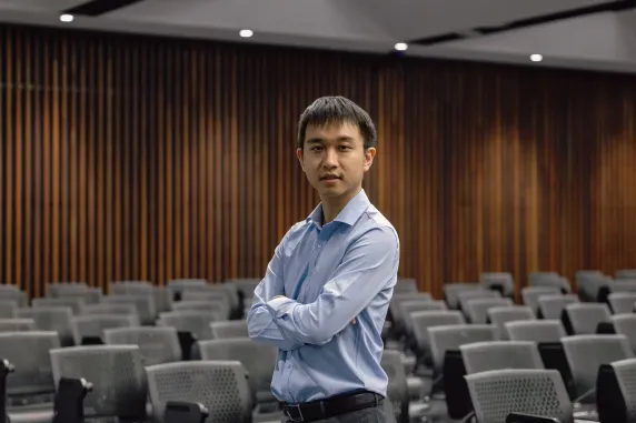 A man in a light blue dress shirt stands with arms folded in the middle of an empty lecture theatre. He faces the camera confidently, with rows of grey chairs behind him and vertical wooden wall panels in the background.