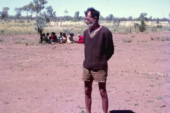 Man standing in a dirt area in the bus, with Indigenous people sitting behind him under a small tree