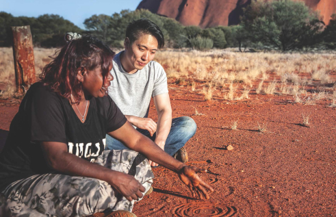 Indigenous woman explaining a figure drawn in the dirt to a man who is sitting with her and listening