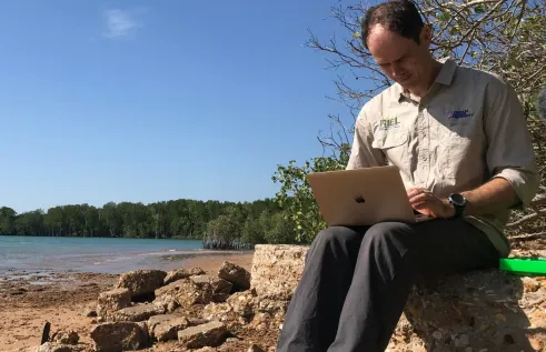 CDU Professor Sam Banks sits on the rocks at the beach with his laptop