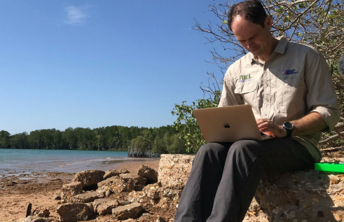 CDU Professor Sam Banks sits on the rocks at the beach with his laptop