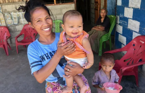 A woman holding a small child with another child standing beside her and an old woman sitting in the background