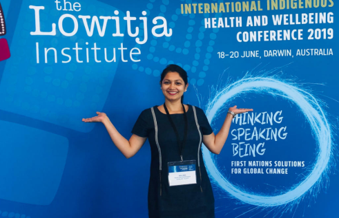Athira smiling in front of a promotion for the International Indigenous health and Wellbeing Conference 2019