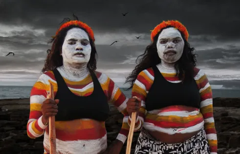 Two woman stand on a beach, with traditional ochre painting on their bodies.