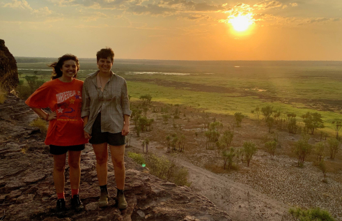 Two people standing on high rock with green plain in background and sun low in sky