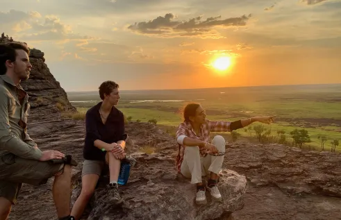 three people on a rock high above a background plain with sun low in sky