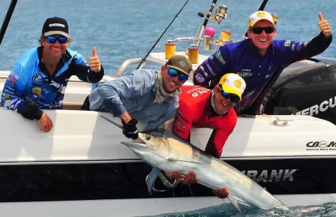 Four men in a boat, two leaning over the side holding a big fish