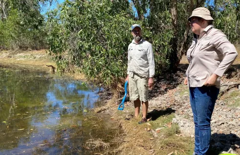 Two people standing on the bank of a water body with trees in the background