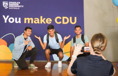 three students posing underneath a sign that says "You make CDU" and a woman taking their photo with a mobile phone