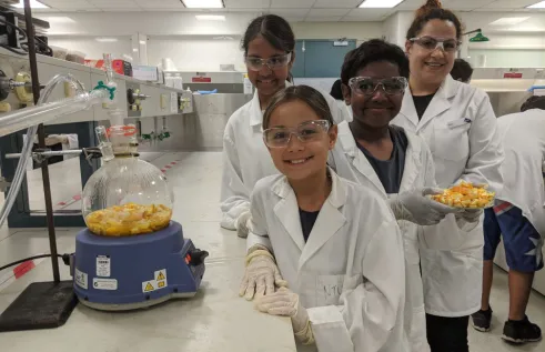 three children and an adult wearing white coats and protective glasses, in a laboratory