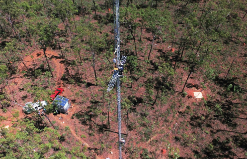 Aerial view of a tall tower with two people working on it. Trees, vehicles, and solar panels far below