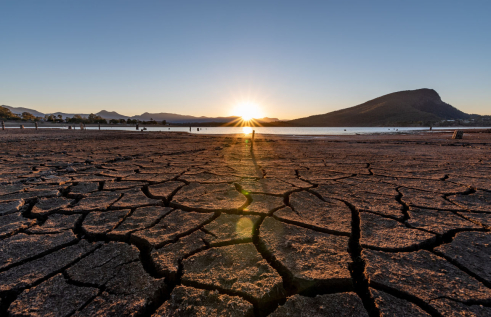 Cracking soil of a dry lake bed, with water in the distance and hills behind. Sun low in the sky behind hills