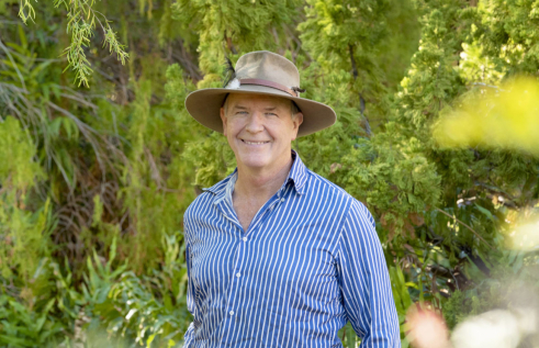Professor Stephen Garnett head and shoulders, wearing blue and white striped shirt and an Akubra hat, with green leafy vegetation in background