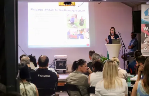 people sitting with backs to camera, Maxine Piggott standing on stage speaking, with presentation slide projected onto screen next to her