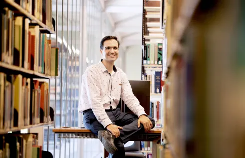 Man sitting on desk surrounded by books in a light filled room.
