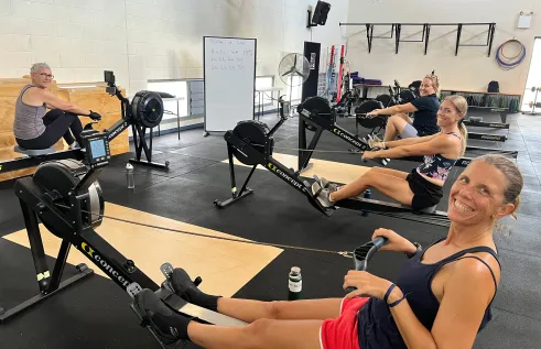 Four women using indoor rowing machines to practice for the upcoming Australian Championships. They are all smiling.