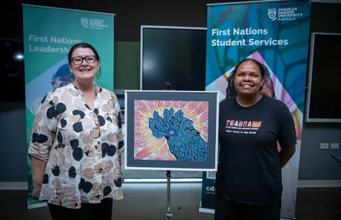 Caucasian woman wearing glasses with a proud smile on her face standing next to an artwork of a blue cockatoo with a young Aboriginal woman on the right also with a big proud smile on her face. 