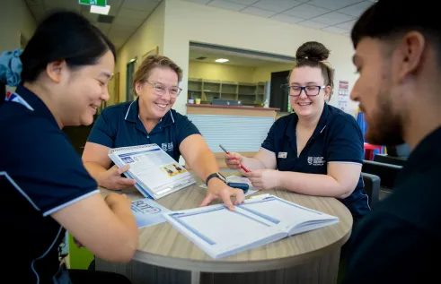Four people sitting around a table. The teacher is smiling while pointing to a book that is sitting on the table in front of the student. 