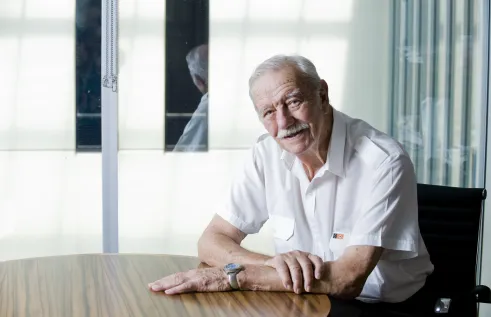 Image of the Honourable Austin Asche AC KC in 2011 sitting at a desk looking straight at the camera. He wears a white shirt with a badge of the Northern Territory flag. 