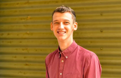 Sam Williams - young man smiling and wearing a light red button up shirt smiling in front of corrugated iron wall