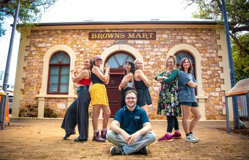 Six women stand behind one man who is seated on the floor while the women look strong and proud standing in poses akin to Charles Angel's.