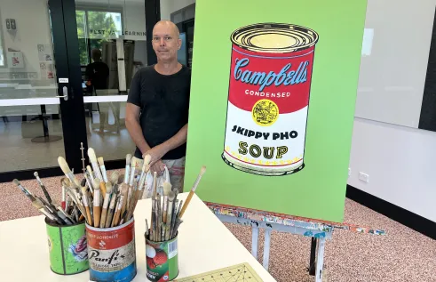 Man in black t-shirt stands next to his artwork called 'Skippy Pho' (2022). On the desk in front of him there are paint brushes in three containers. 