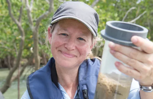 Julia Fortune holding a clear container with brown water and mud inside, with water and mangroves in the background