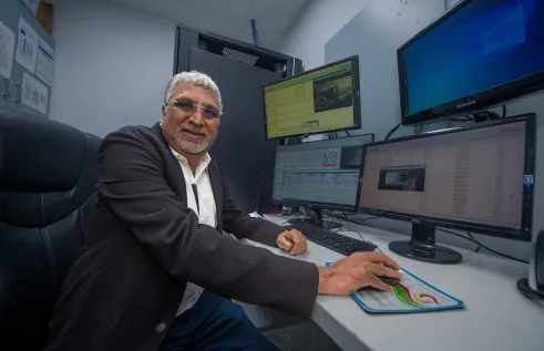 Man sitting at desk with computer screens in front of him. He is holding the mouse but looking directly at the camera and smiling. 