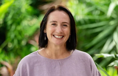 Caucasian women with brown hair smiles widely at the camera. She is outside in front of some green foliage and is wearing a lilac coloured top. 