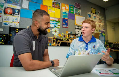Two people sitting at a desk. A laptop is open in the middle. The University student on the left smiles at the Year 9 student on the right. They're in an animated discussion. 