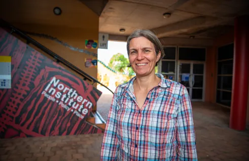 Caucasian woman with grey hair and a plaid blouse standing in front of a stairway that says Northern Institute. 