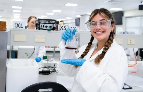 A young woman wears a white lab coat and is holding a glass beaker inside a lab. She has a wide smile. 