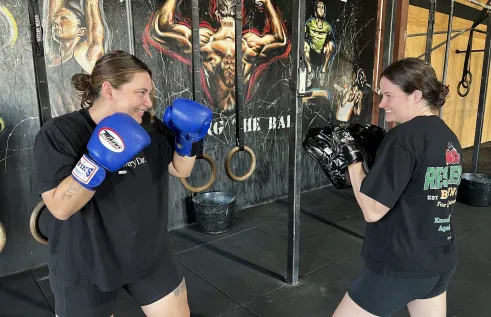 Two women wearing black fitness gear and boxing pads on their hands stand in a mock-spar stance. They both have smiles on their faces. 