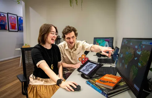 Young woman and man sit at a desk looking animatedly at a computer screen. The young man is pointing. The young woman has a bright smile on her face. 