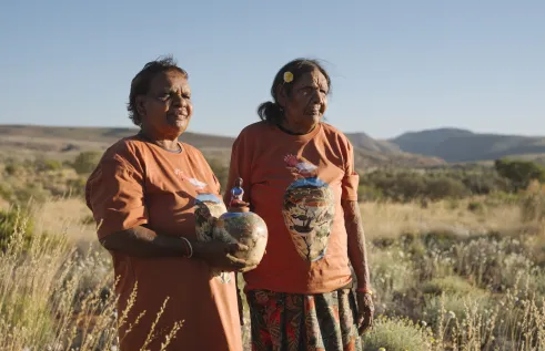 Two First Nations women stand outside surrounded by grass and mountains. One is holding a clay pot with drawings on it. They both look out into the distance.
